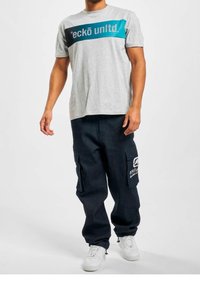 Gray t-shirt featuring a black and teal logo band across the chest, paired with dark cargo pants and white sneakers, showcasing large side pockets.