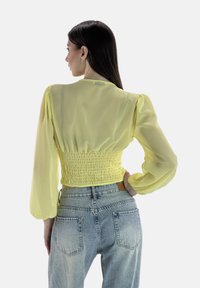Gelbe durchsichtige Bluse mit Puffärmeln und einem gerafften Rücken, kombiniert mit hochgeschnittenen hellblauen Jeans. Der Stoff ist leicht und fließend.