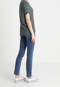Kvinde iført en mørkegrøn mønstret kortærmet top, blå slim jeans og hvide platform sneakers, der står sidelæns mod en ensfarvet baggrund.