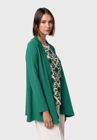 Grön, lätt cardigan med öppen frontdesign, bärs över en mönstrad topp med mörkgröna blommönster, parat med vita byxor.