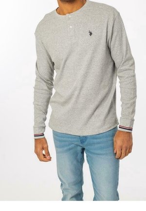 Uomo che indossa una maglia henley a maniche lunghe grigio chiaro con logo nero sul petto e jeans blu, in piedi con le mani rilassate lungo i fianchi.