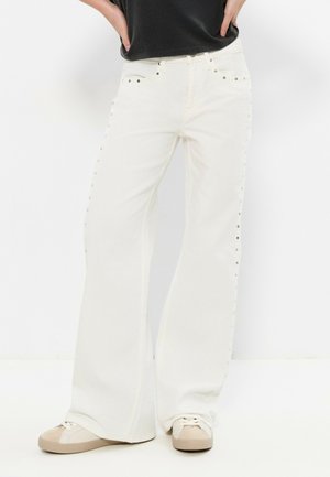 Personne portant un jean blanc à jambes larges avec des clous sur les côtés, associé à des baskets beige et blanches et un haut noir, debout les mains sur les hanches.