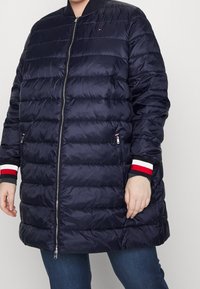 Tmavě modrá prošívaná péřová bunda s vodorovným prošíváním, s předním zipem, bočními kapsami a pruhovanými červenými, bílými a modrými ozdobami na manžetách.