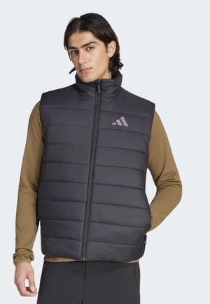 Man draagt een zwart, gevoerd mouwloos vest over een bruin shirt met lange mouwen, handen ontspannen, staat tegen een effen lichte achtergrond.