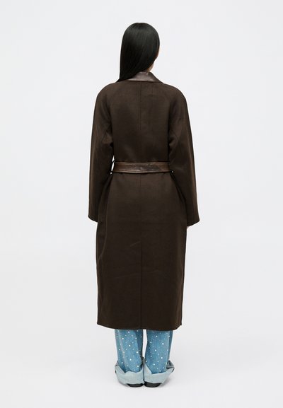 Άτομο με μακριά μαύρα μαλλιά στέκεται με την πλάτη στραμμένη, φορώντας ένα μακρύ καφέ παλτό με δερμάτινη ζώνη και μπλε τζιν με πουά μανσέτες.