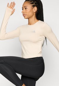 Μπεζ αθλητικό τοπ με μακριά μανίκια, εφαρμοστή σχεδίαση και τρύπες για τον αντίχειρα, σε συνδυασμό με μαύρο κολάν ψηλόμεσο με λεπτομέρειες αντιθετικής ραφής.