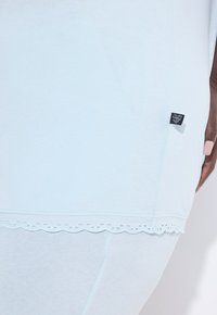Svetle modré bavlněné tričko s krajkovým detaily na lemu ve tvaru obloučku. Obsahuje malou textilní štítek na boku, která podtrhuje jemnou strukturu a neformální design.