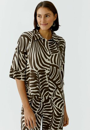 Femme portant une chemise boutonnée marron et blanche à motifs zébrés abstraits avec un pantalon assorti, souriante et regardant sur le côté.