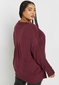 Burgunderfarbener Oversized-Pullover mit gerippter Textur, überschnittenen Schultern, seitlichen Schlitzen und Knopf-Akzenten an den Ärmeln. Aus weichem Strickstoff.