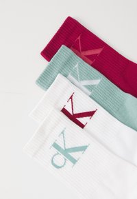 Four pairs of socks in red, light teal, white, and light gray. Each has a ribbed texture and a large, contrasting logo on the cuff.