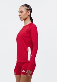 Crvena atletska majica dugih rukava s okruglim izrezom i bijelim trostrukim trakama. U paru s crvenim uskim šortsevima s Adidas logotipom.