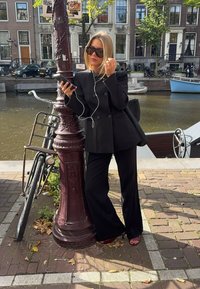 Svart skräddarsydd dubbelknäppt kavaj och matchande vida byxor, parat med öppna klackskor, stående bredvid en cykel och en kanal.