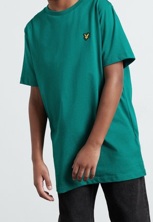 Barn i en ensfarvet grøn T-shirt med korte ærmer og en lille gul fuglelogo på brystet, kombineret med mørke bukser.