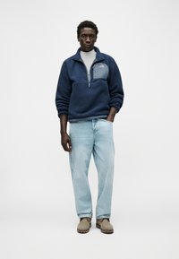 Άνδρας που στέκεται φορώντας μπλε ναυτικό φλίς πουλόβερ, λευκό ζιβάγκο, ανοιχτό μπλε τζιν και μπεζ μπότες πάνω σε απλό φωτεινό φόντο.