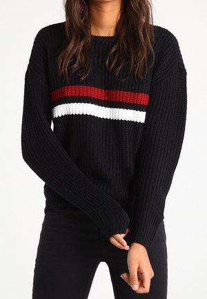 Personne portant un pull en maille noir avec des rayures horizontales rouges et blanches sur la poitrine, associé à un pantalon noir.