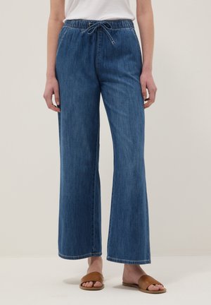 Frau trägt hoch taillierte blaue weite Jeans mit Kordelzug und braunen Schlupfsandalen, steht vor einem schlichten hellen Hintergrund.