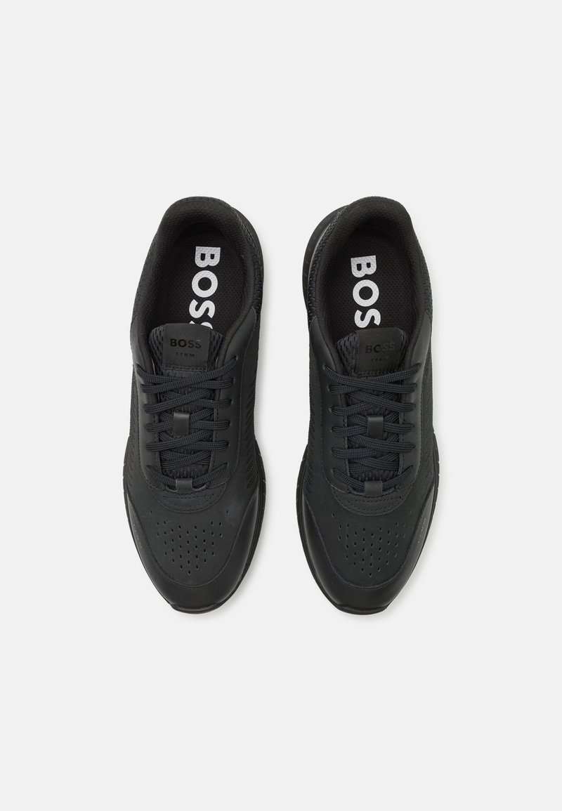 Zapatillas deportivas negras con parte superior de piel lisa y malla, que presentan perforaciones, cordones redondos y un logotipo prominente en las plantillas.