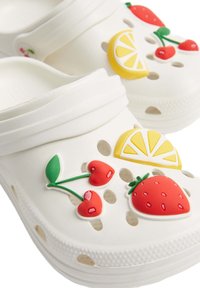 Des sabots blancs à enfiler avec des trous de ventilation, décorés de cerises rouges, d'une fraise rouge et d'une tranche de citron jaune.