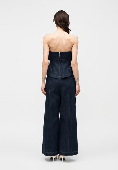 Hosszú, sötét hajú nő, aki ujjatlan, sötét farmer anyagú, vállpánt nélküli felsőt visel hátul cipzárral, és hozzáillő, bőszárú nadrágot, fehér magas sarkú cipővel.