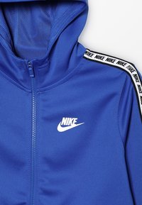 Sudadera con capucha con cremallera de color azul de Nike, hecha de tela suave, con capucha forrada de malla, cierre de cremallera y cinta con logotipo en blanco y negro a lo largo de los hombros.