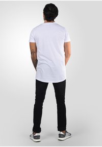 Camiseta blanca de manga corta con un dobladillo curvado, combinada con vaqueros negros y zapatillas a rayas grises, vista desde atrás.