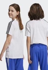 Camiseta blanca de algodón con rayas negras en los hombros, combinada con pantalones cortos deportivos azules. Diseño sencillo con mangas cortas y ajuste relajado.