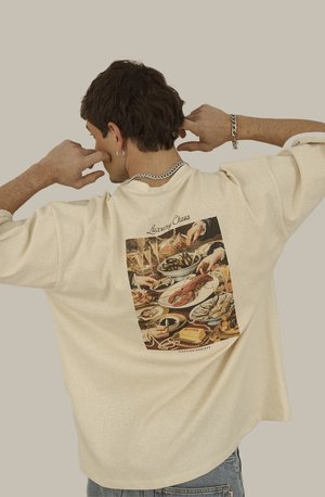 Camiseta oversized color crema con un gráfico colorido de temática gastronómica en la parte posterior, acompañado del texto "Luxury Chaos" encima. Textura pesada, diseño casual.