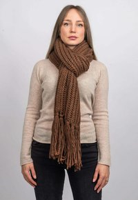 Sciarpa lavorata a maglia marrone con motivo testurizzato e frange alle estremità, indossata sopra un top beige chiaro, posizionata su uno sfondo neutro.