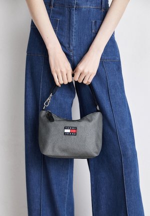 Person wearing blue denim overalls holding a small gray Tommy Jeans handbag by its black strap in front of their body.