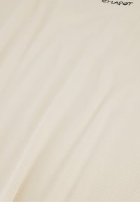 Beige, gestructureerde stof met subtiele verticale strepen; heeft geborduurde merknaam in de bovenhoek. Glad en zacht aan de aanraking.