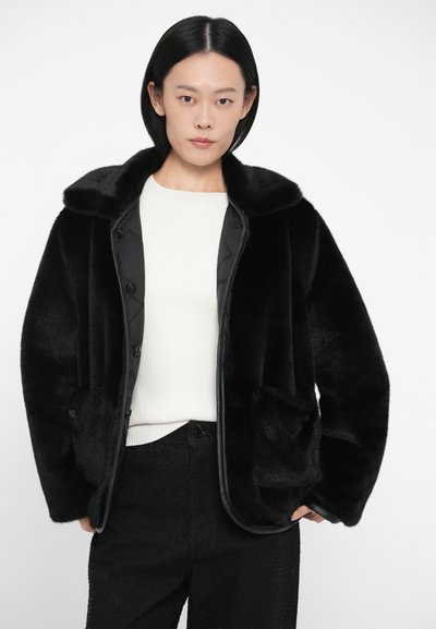 Μαύρο ψεύτικο γούνα μπουφάν με φαρδύ γιακά, κουμπί κλείσιμο και πλαϊνές τσέπες. Διαθέτει γυαλιστερή υφή και απαλή υφή. Φορεμένο πάνω από ένα λευκό μπλουζάκι.