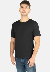 Schwarzes Kurzarm-T-Shirt aus reinem Baumwollstoff, mit Rundhalsausschnitt und einem dezenten Logo-Detail auf der Brust. Kombiniert mit blauen Jeans.