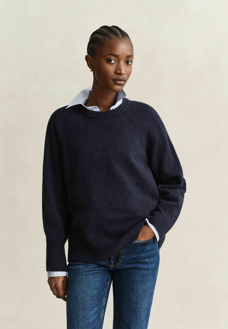 Femme aux cheveux tressés portant un pull bleu marine foncé sur une chemise blanche à col et un jean bleu, debout avec une main dans la poche.