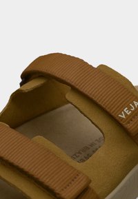 Καφέ vegan σανδάλια με υφασμάτινα λουριά, διαθέτουν επενδεδυμένη, μπεζ σόλα και λεπτομερές ράψιμο. Το λογότυπο της μάρκας είναι ορατό στο λουρί.