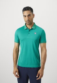 Muž v tyrkysovom polo tričku s krátkym rukávom a malým logom na ľavej hrudi, v tmavo námorníckych nohaviciach, stojí proti jednoduchému pozadiu.