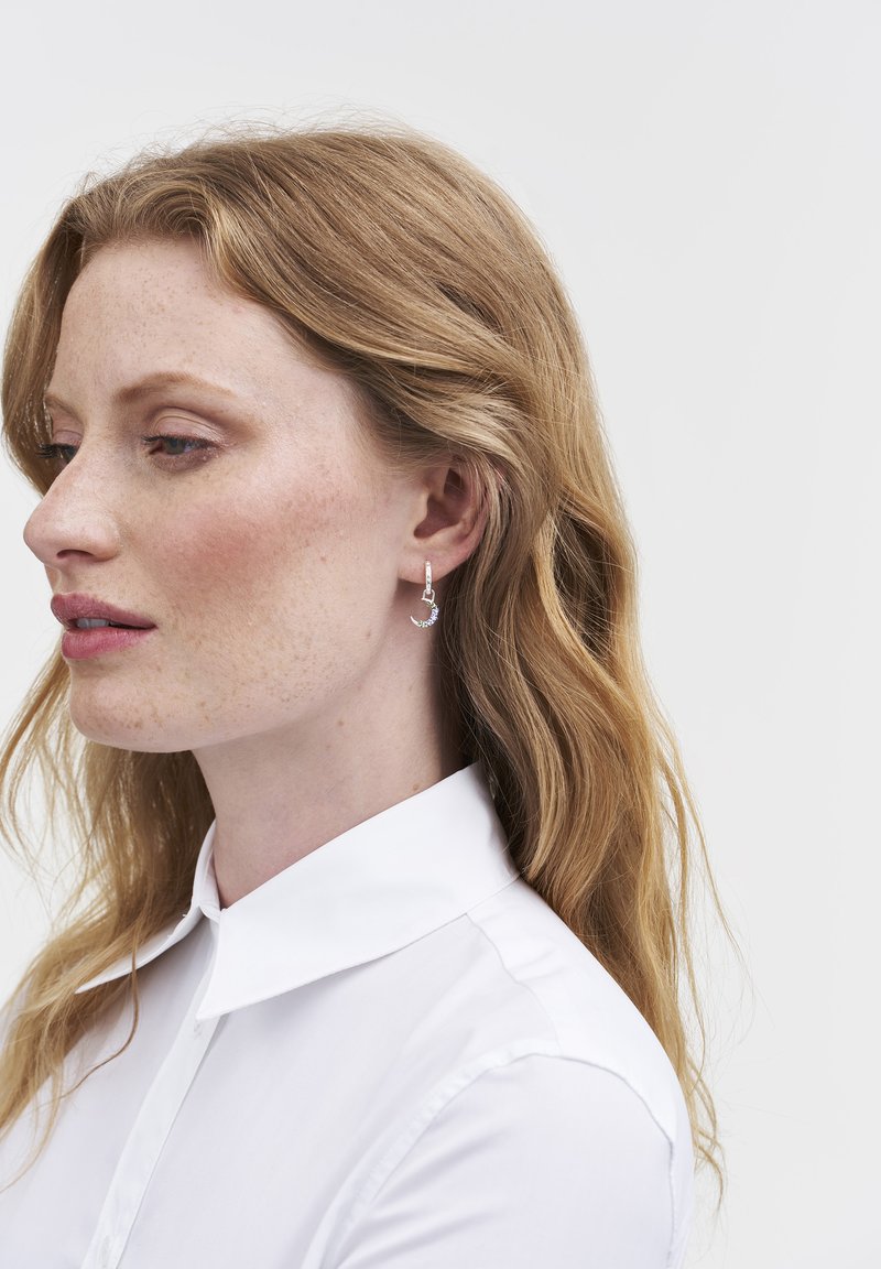 Pendientes de aro de plata con un pequeño colgante y acentos brillantes, usados por una persona con cabello ondulado, vestida con una camisa blanca de cuello.