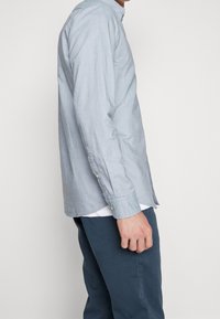 Ljusblå skjorta med knappar och långa ärmar, knäppningar vid ärmarna och rak skärning, tillsammans med marinblå byxor. Mjuk tygstruktur, minimal detaljer.