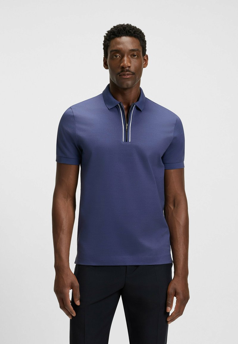 Granatowa koszulka polo z krótkim rękawem, z kołnierzykiem stojącym i pół-zamkiem z przodu, z białymi akcentami wzdłuż zamka. Gładka faktura materiału.