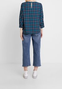 Chemise à carreaux en teal et marine, avec une fente dans le dos et des manches trois-quarts, associée à un jean large bleu clair et des baskets blanches.