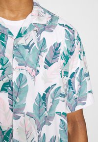 Kortärmad skjorta med vit bas som har ett livligt mönster av gröna och rosa blad, knäppt framtill och med en camp krage design.