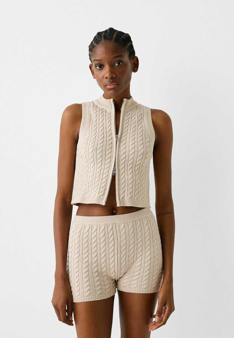 Gilet corto lavorato a maglia beige con colletto alto e chiusura con cerniera, abbinato a pantaloni corti a vita alta in maglia abbinati con motivo a treccia.