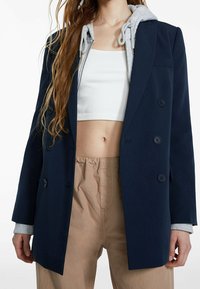 Blazer doppiopetto blu navy con colletto a scollo, spalline, tre bottoni e felpa grigia con cappuccio sotto. Indossato con pantaloni beige.