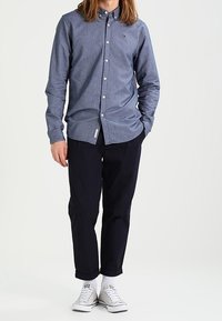 Camisa de botones en tela de chambray azul oscuro, con un pequeño logotipo en el pecho, combinada con pantalones oscuros y zapatillas blancas.