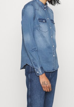 Person iført en falmet blå denimskjorte og blå jeans med en tatovering af flammedesigns på venstre hånd.