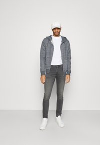 Γκρι φούτερ με φερμουάρ και κουκούλα, λευκό T-shirt από κάτω, γκρι skinny παντελόνι και λευκά αθλητικά παπούτσια. Λείο ύφασμα με χαλαρό σχέδιο.