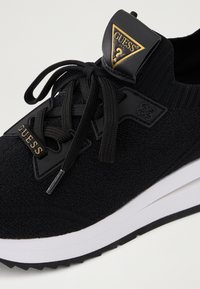 Melnas auduma sporta kedas ar adītu augšdaļu. Iekļauts melns gumijas logotipa uzlīme un teksturēta saite, papildināta ar baltu starpsoļu un melnu zoli.