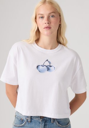 Ξανθιά γυναίκα που φοράει άσπρο κοντό T-shirt με μπλε σχέδιο κερασιού και μπλε τζιν, πάνω σε απλό φόντο.