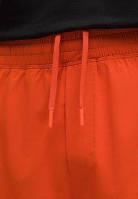 Nahaufnahme von leuchtend orangefarbenen, elastischen Hosen mit passendem Kordelzug und roten Nähdetails vor einem schwarzen Hintergrund.