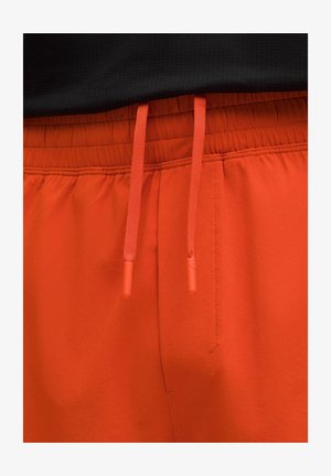 Lähedal vaatamine erkoranžidest elastsetest vöökohtpükstest koos sobivate paeladega ja punase õmbluse detailidega musta tausta ees.