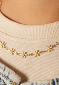 Gouden ketting met een ontwerp van witte en gele madeliefjes, geplaatst tegen een gestructureerde beige stof en blauwe denim.